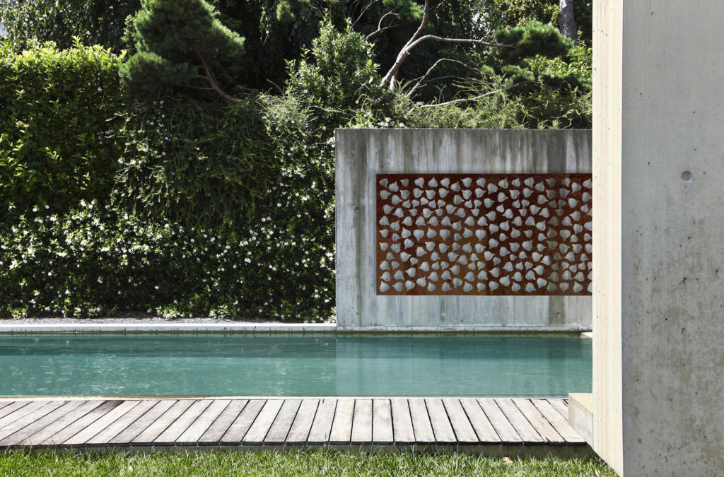 Decorative Screen made from corten steel with natural rusted finish hanging on concrete wall over pool in modern landscape