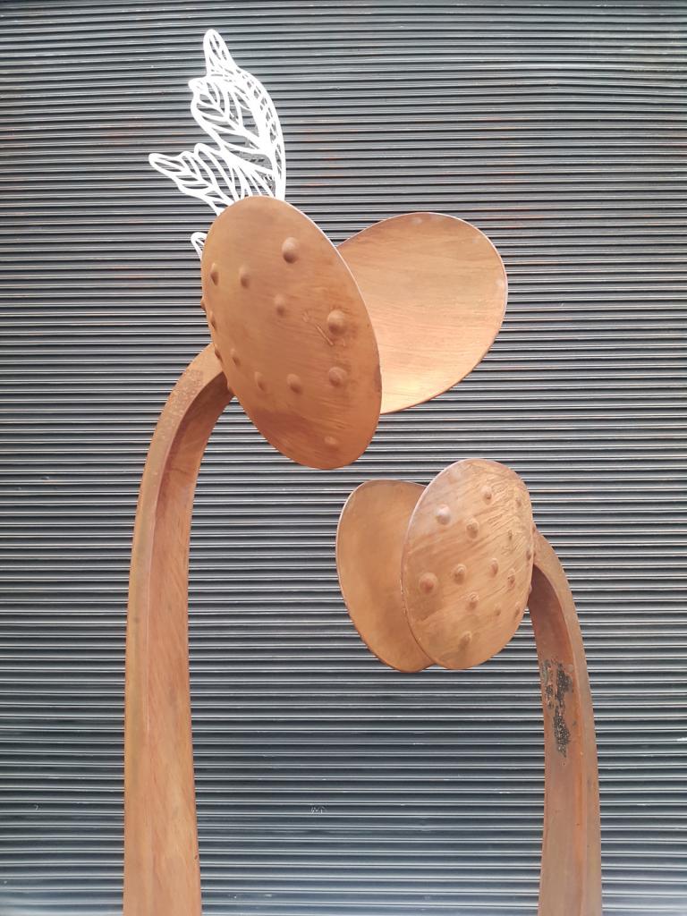 Tall modern sculpture representing a new seedling featuring a stainless-steel leaf emerging from the center of the main seedling. Corten steel with a natural rusted finish.