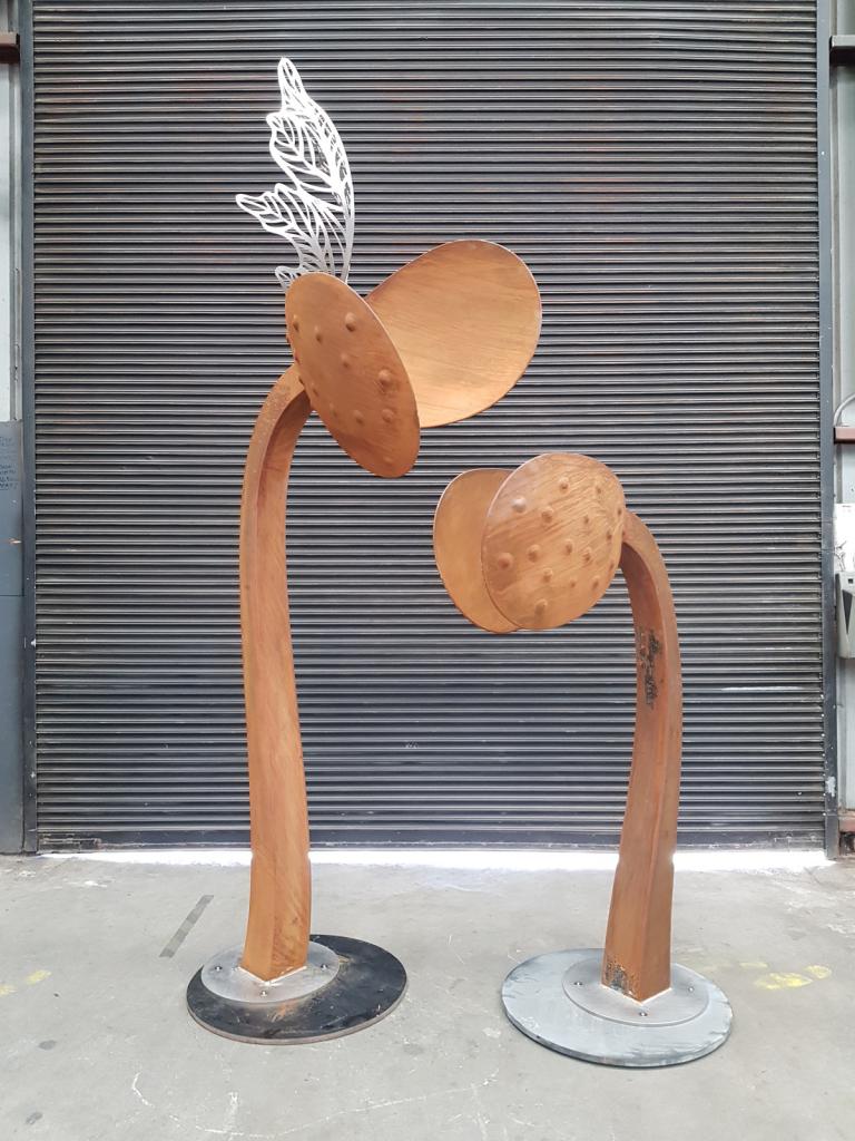 Tall modern sculpture representing a new seedling featuring a stainless-steel leaf emerging from the center of the main seedling. Corten steel with a natural rusted finish.